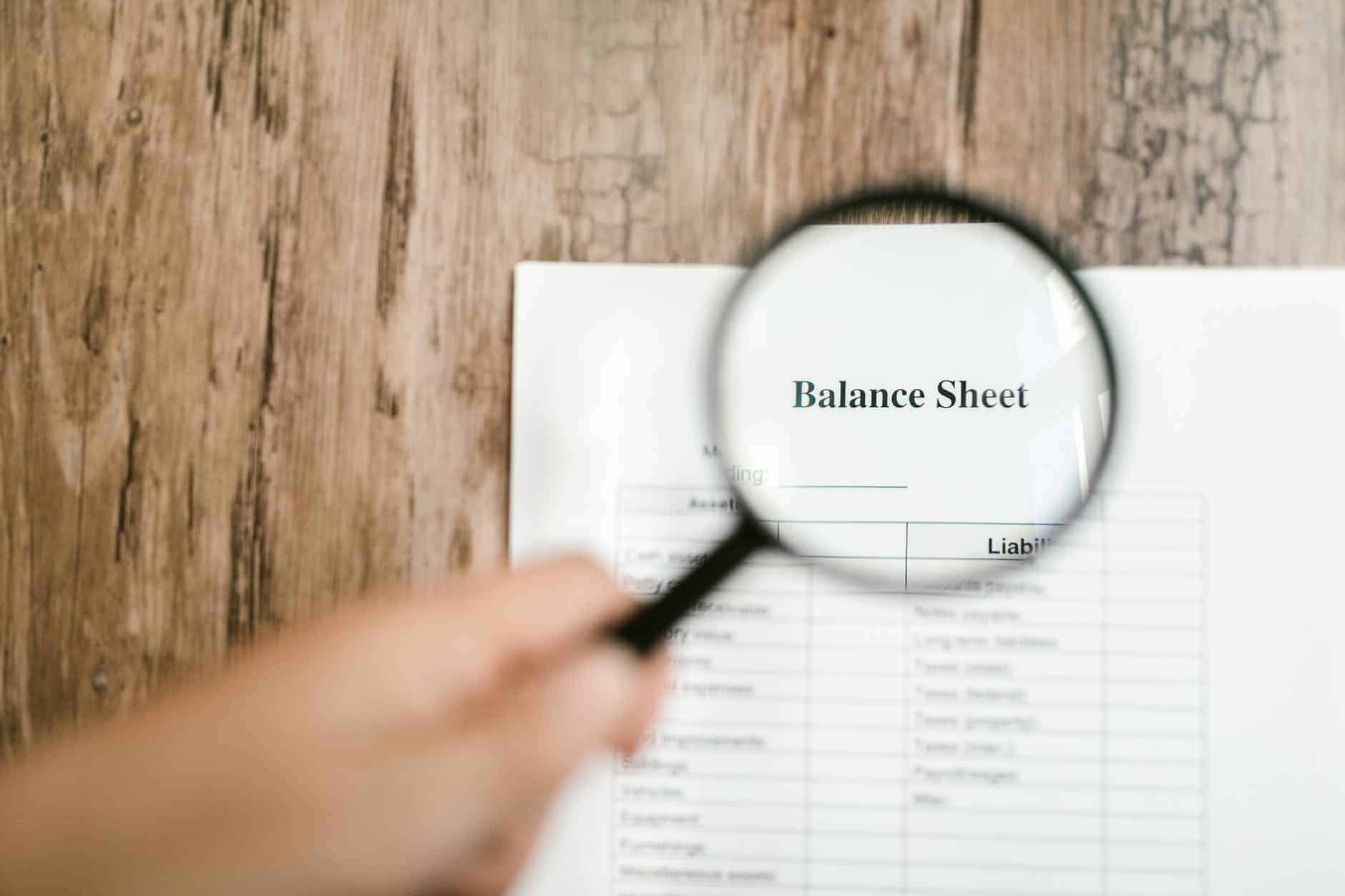 hand holding a magnifying lens focusing in on a piece of paper that says balance sheet