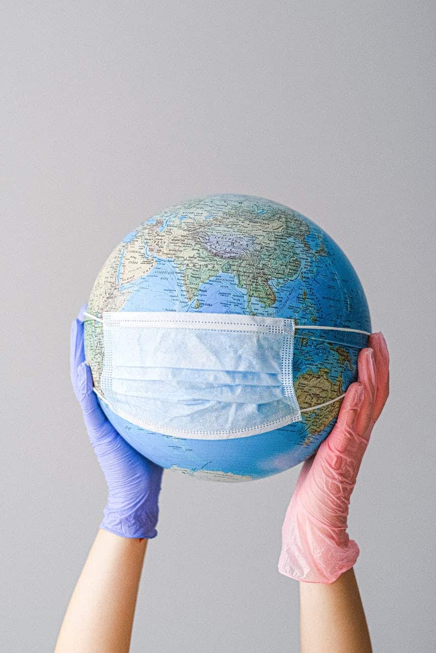 Two hands holding up a globe of the earth with a face mask on it