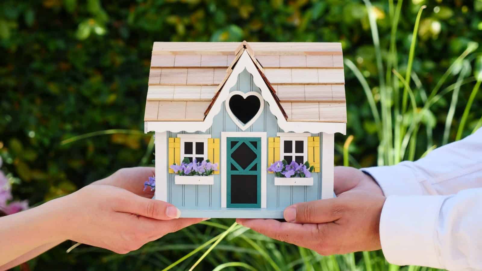 Two pairs of hands holding a small wooden house in their palms