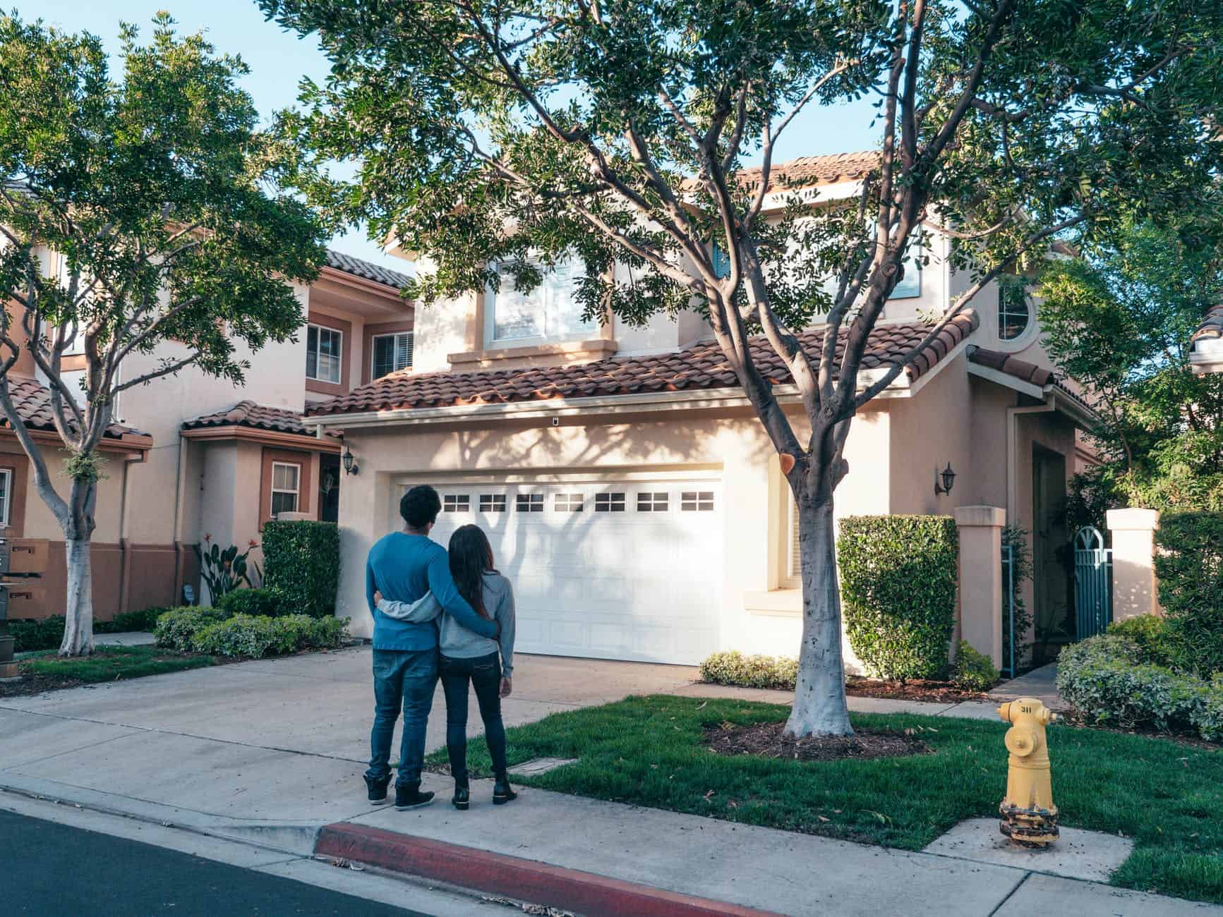 two people standing on a sidewalk holding each other while looking at the front of a house that has a tree in the small front yard next to the driveway