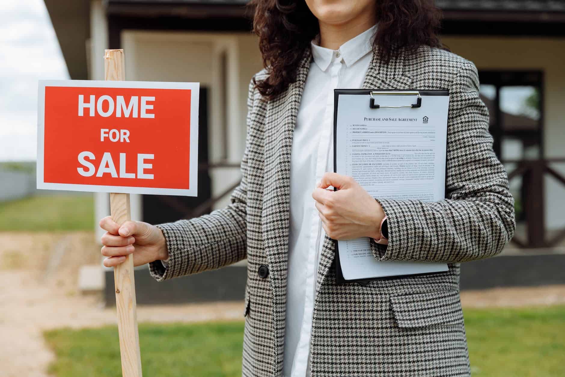 Lady in a suit holding a clipboard in left hand and a home fore sale sign in right hand