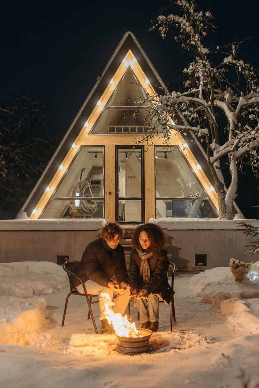 a man and a woman sitting by a fire in the snow with a house with lights on behind them