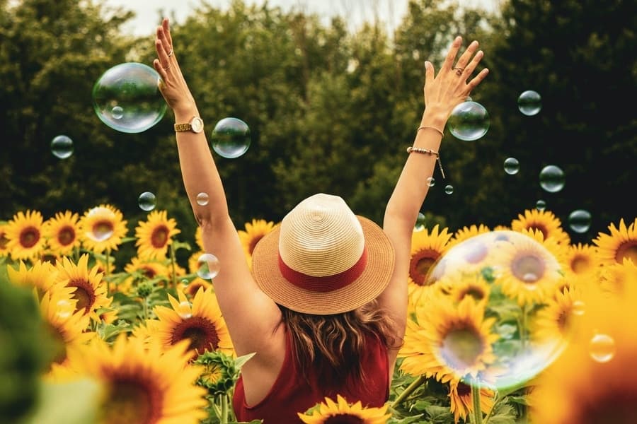 woman wearing a hat raising her arms up while standing in a field of sunflowers with bubbles floating in the air